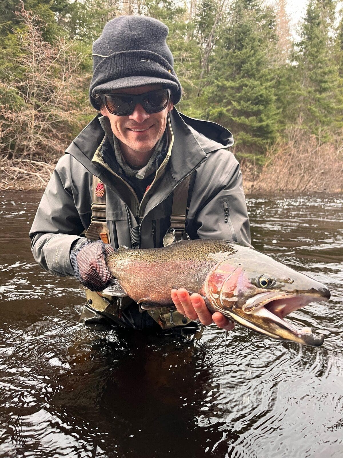 An angler in a wool hat holding a large male steelhead with a hooked jaw over the water